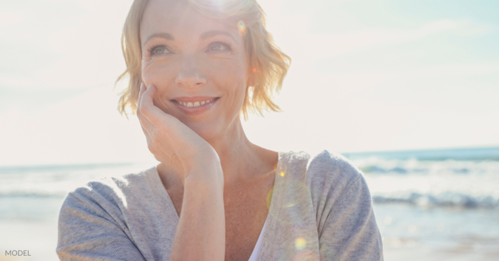 Woman (model) holds a hand up to her face while smiling on the beach.