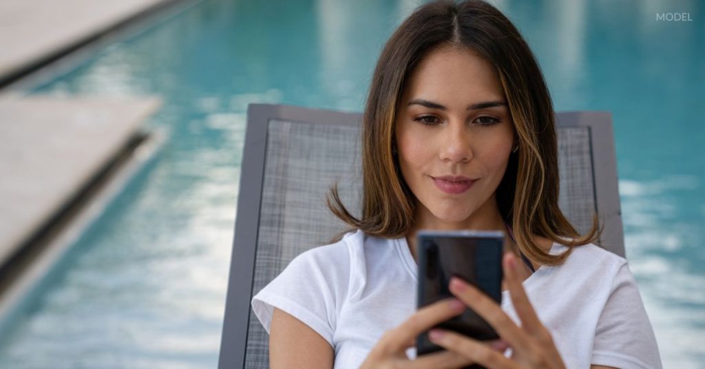 Woman (model) scrolling on her phone by the pool.