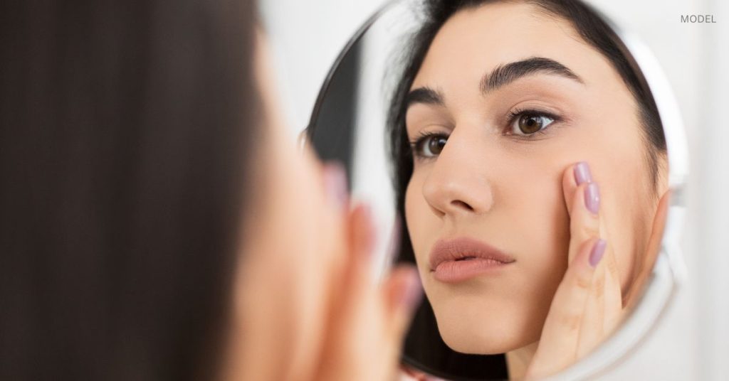 Woman (model) inspecting her skin while looking in the mirror.